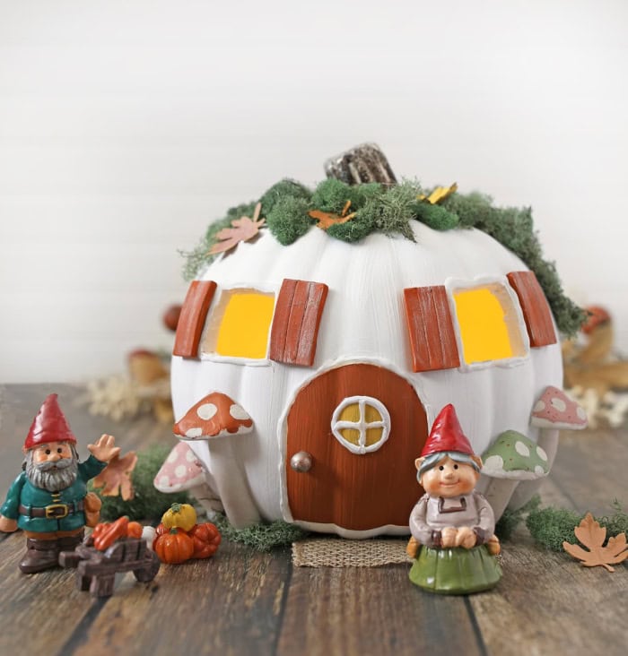A white pumpkin decorated to look like a fairy cottage.  The inside is carved out for lights, it has two windows a green moss roof and is surrounded by little elf characters.