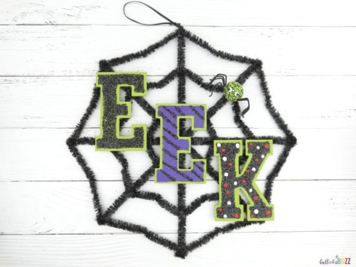A spider web shaped wreath with EEK written on front.  Tje wreath is black, brigjt green and purple withh sparkles.