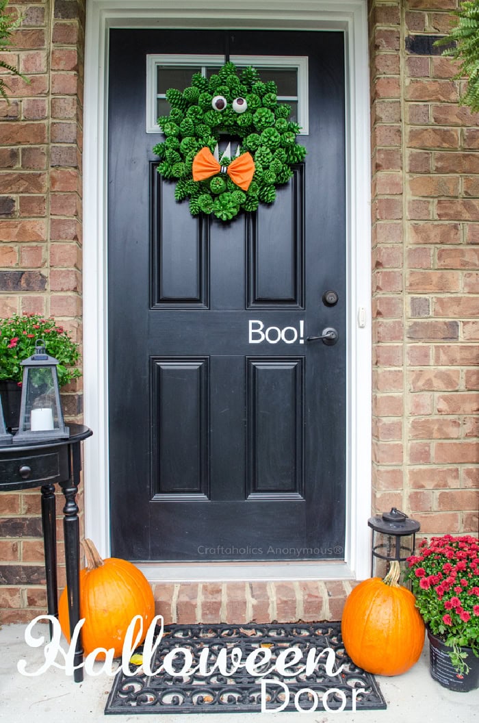 A black front door decorated for Halloween with a cute DIY Halloween wreath made with green pinecones and eyes to look like a fun monster.