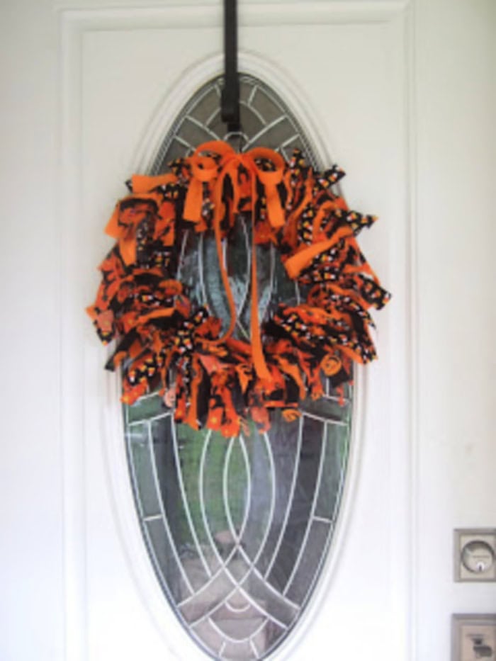 A bright orang and black Halloween printed fabric made into a cheerful and fun Halloween rag wrath.  The bright wreath is hanging on a white front door.