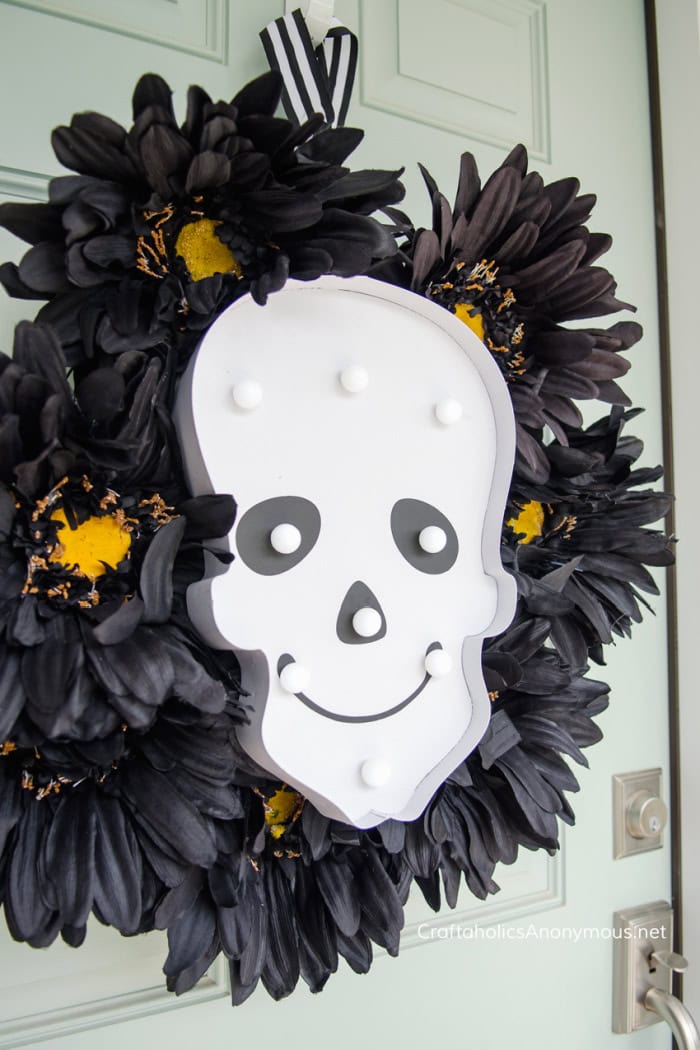 A Halloween wreath made with black flowers arounnd the outside and a finny skull face marquis sign in the center for decoration.