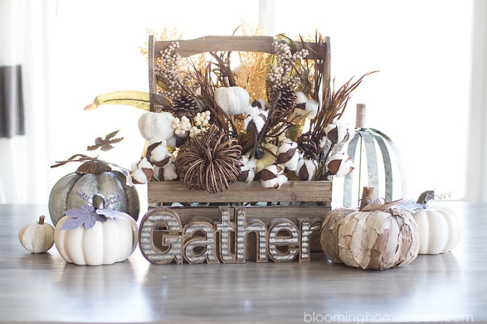 A diy pumpkin centerpiece in a wood basket.  The wood basket is filled with greenery and surrounded with neutral colored pumpkins.