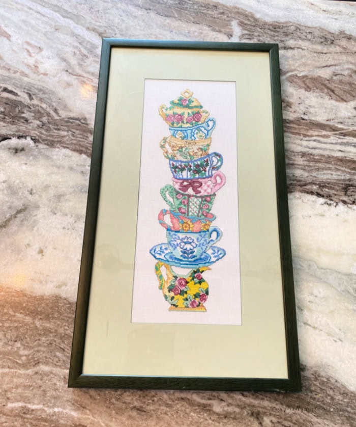 The before picture of a teacup needle point vintage find. It has a stack of colorful teacups a soft green mat and a dated green frame.