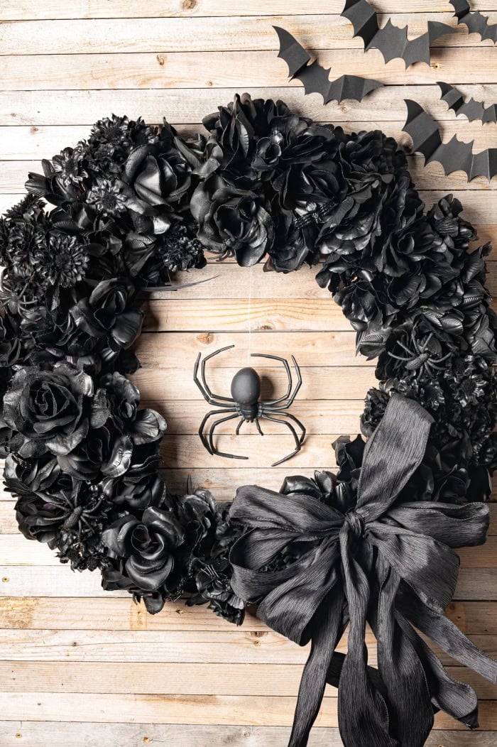 A all black Halloween wreath made with flowers, painted black then decorated with a elegant block ribbon bow.  There is a spider in the center.
