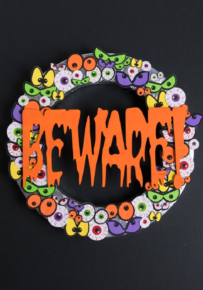 A very colorful and bright Halloween wreath DIY with assorted eyeballs for the background.  The centerpiece is  the word Beware inb right orange.