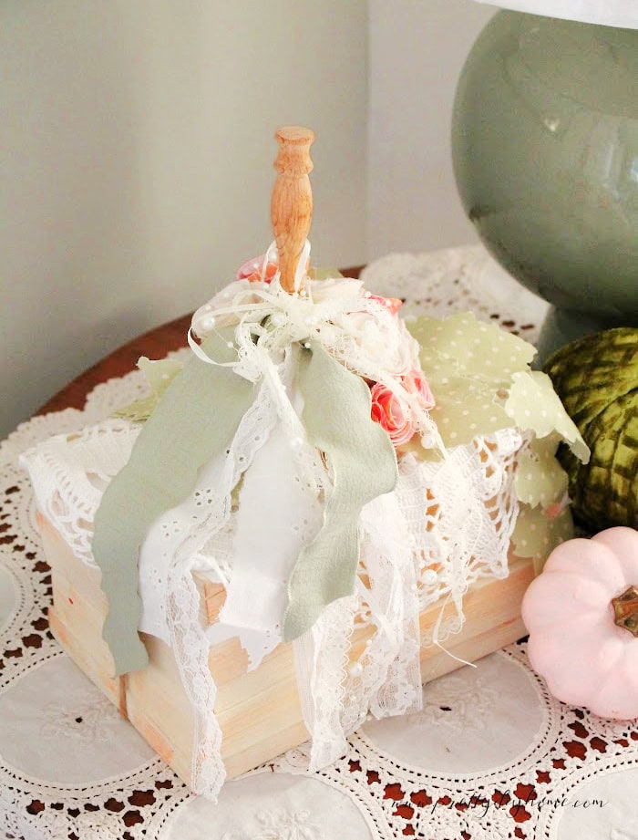 A DIY bookstack pumpkin with assorted fabric ribbons and vintage lace cascading down the side. It is sitting on a lace tablecloth with a green lamp in the background and a pink pumpkin nestled in beside it.
