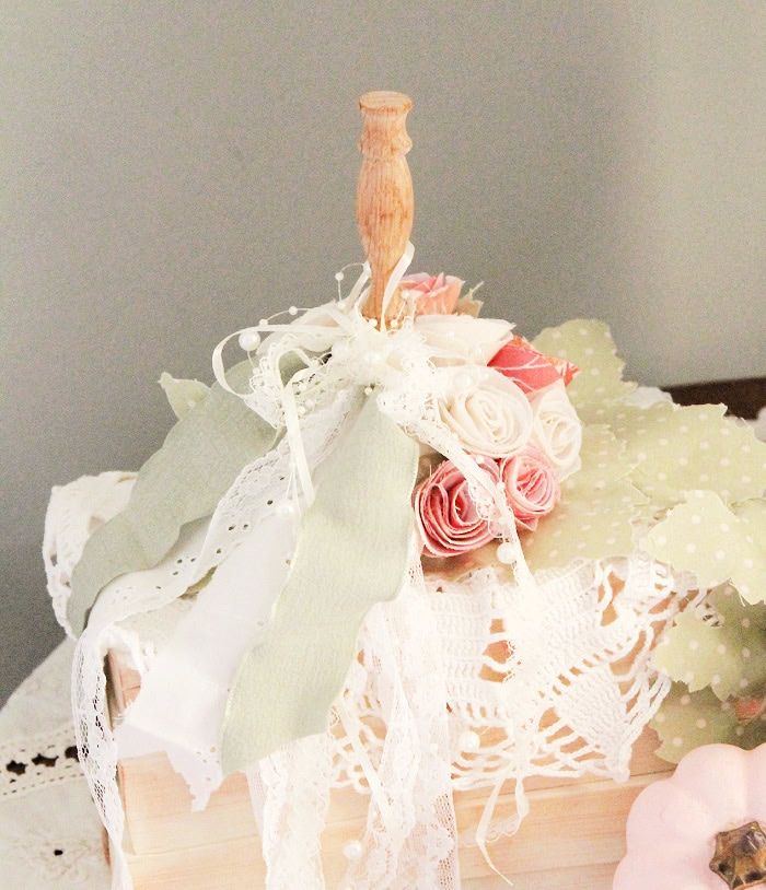 A fall pumpkin craft in neutral colors for Fall decor. The pumpkin craft is made with vintage lace ribbons on one side, handmade rolled roses on the other in pinks, whites, and creams, it has a neutral wood stem, and the pumpkin is made using books.