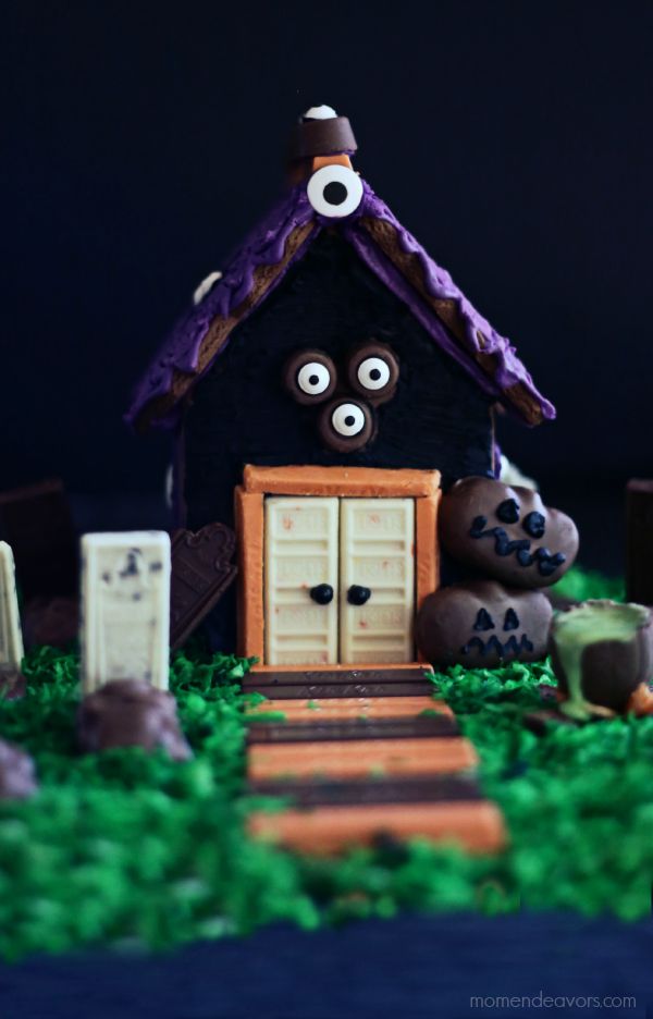 A haunted Halloween gingerbread house with cute candy eyes, a purple rought, candy bar sidewalk and two jackolantern cookies vby the front door.