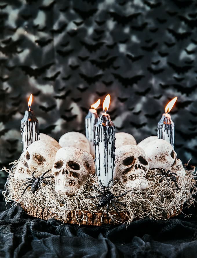 A scary gothic styles centerpiece for Halloween. The centerpiece is on a large log round with white grass, several white skulls, spiders, and white candles with black tears.