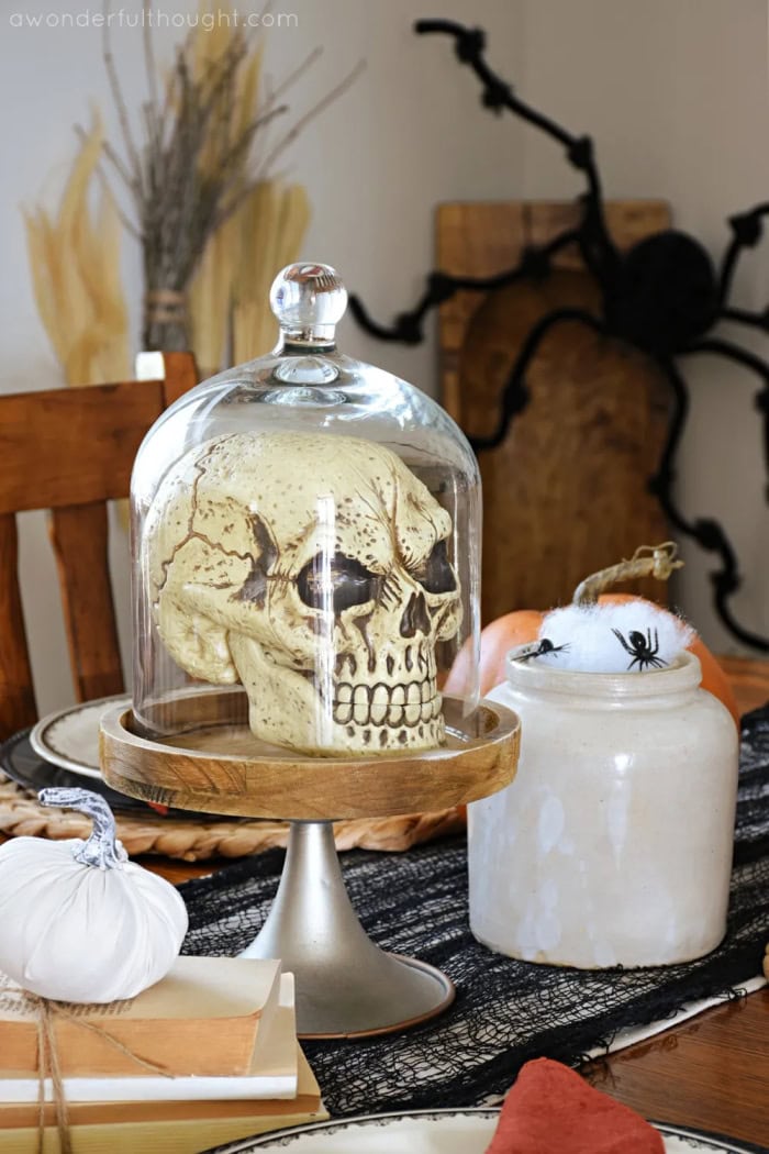 A Halloween tablesetting in soft golds, browns and oranges with a skull themed centerpiece in a cloche.  Their is a giant Halloween spider in the background on the wall.