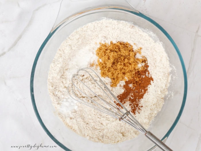 Mixing ingredients for a pumpkin spice pancake recipe in a clear glass bowl with a wisk. You can see pumpkin spice, and cinnamon, and brown sugar on top of the flour.