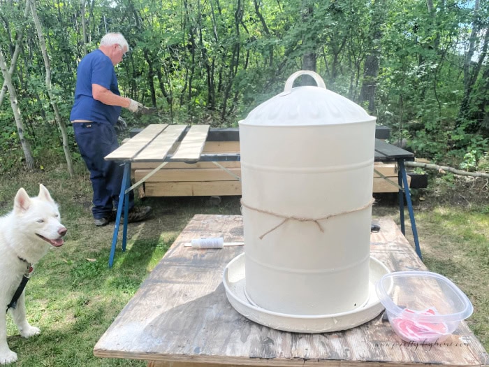 A large metal chicken feeder being painted in an outdoors work area with spray paint.