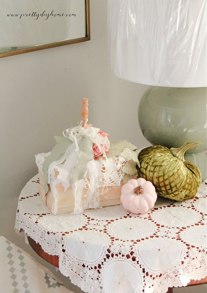 A Vintage Pumpkin craft made with a vintage handmade doily and a bookstack in soft pastel colors for Fall. The diy pumpkin is sitting on a table with lace tablecloth assorted velvet pumpkins and a green lamp.