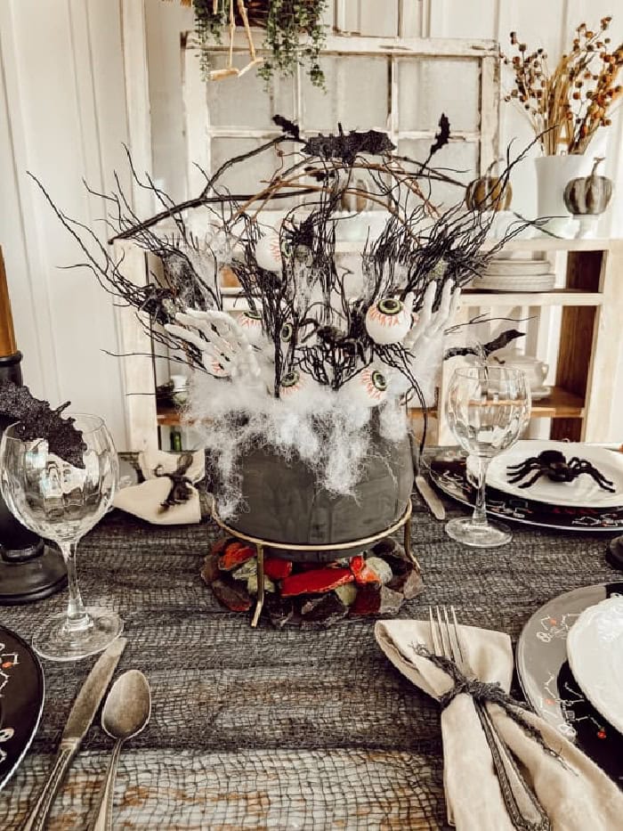 An elaborate witches brew Halloween centerpiece in the center of a tablesetting.  The centerprice is made in a witches cauldron over faux coals and is filled with branches, bats, eyeballs and  wispy cotton to resemble smoke.