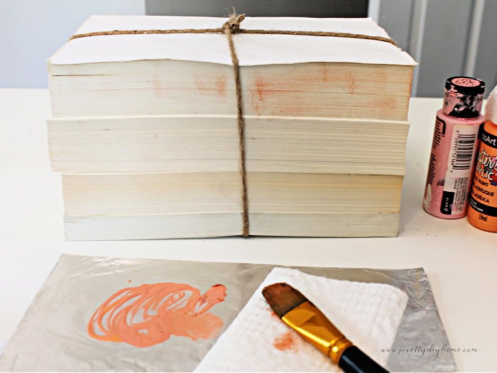 A stack of books put together to as a pumpkin diy. The sides are being dry brush painted with a pinkish coral paint.