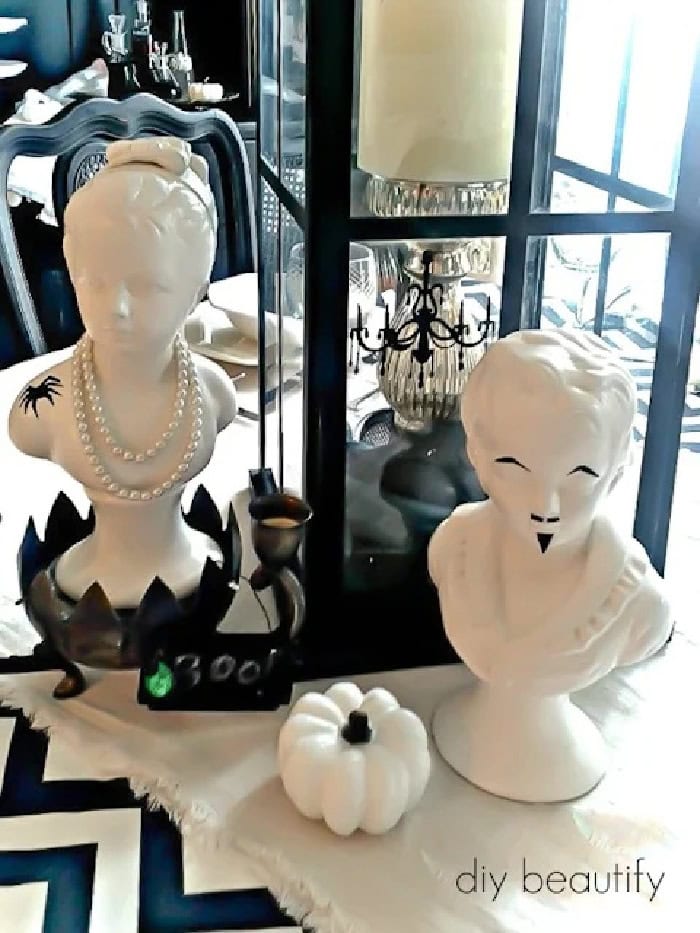 A Halloween centerpiece consisting of two all white heads busks and a pumpkin. The centerpiece is both elegant and spooky.