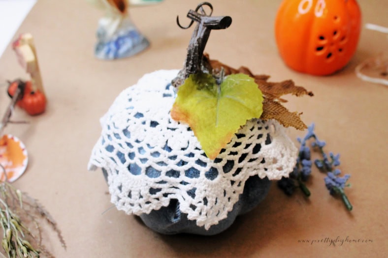 Adding leaves to a blue velvet Dollar Tree pumpkin, the pumpkin is covered with a vintage doily and there are small blue flowers beside it.