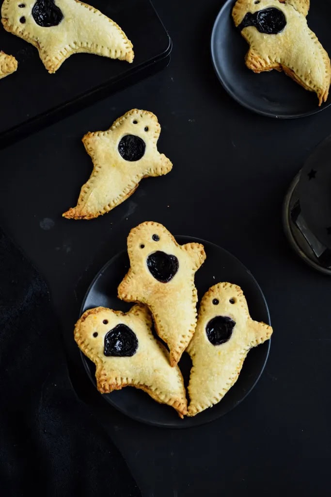 Six boo berry hand pies that look like little ghosts.  Each pie is in a ghost shape with little eye and a large mouth.  They have a golden brown crust and blueberry filling.