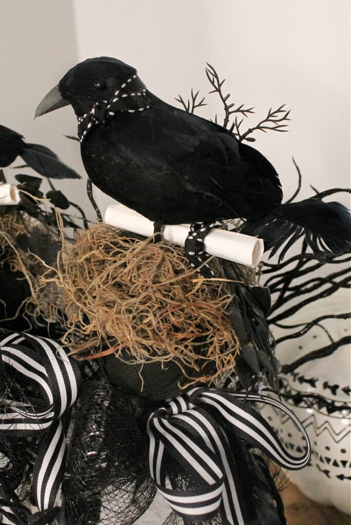 The top of a neutral witch wand Halloween decoration with a black raven, gold raffia, and black and white lace and then striped ribbon and bows.
