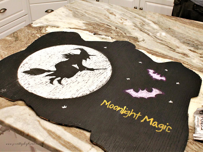 A finished Halloween chalkboard art with a witch silhouette in a white full moon, two purple bats and gold letters that say Moonlight magic. The art is done, but the frame needs to be added to the chalkboard.