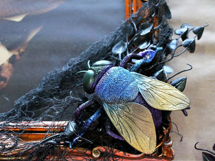 A large plastic and colorful house fly is attached to the bottom corner of a Halloween frame like a little snack.