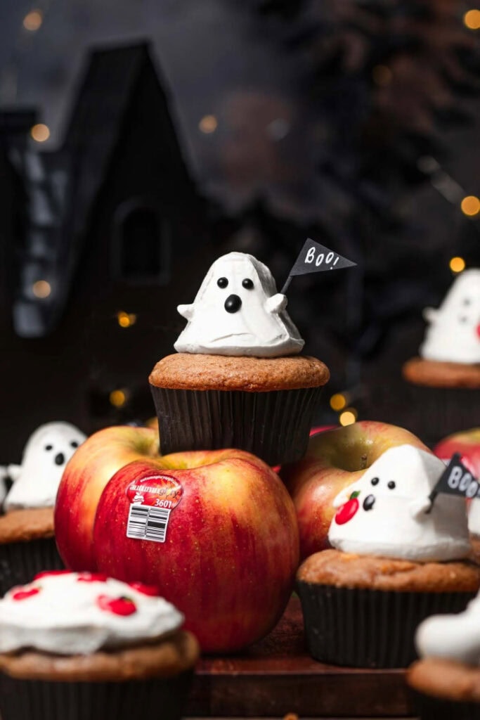 A pile of white ghost cupcakes stacked with apples.  Its a very dark and moody image so the little ghosts stand out.