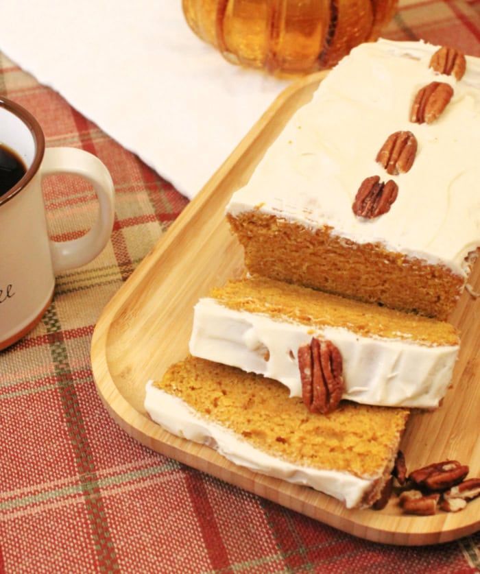 The best pumpkin bread recipe with a whole loaf sitting on a small wood tray. The pumpkin bread is decorated with white sour cream glaze and pecans down the center, and is being served on a white plate.