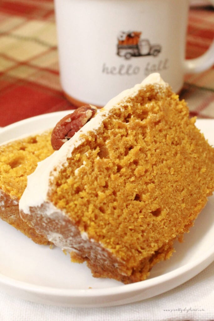 Pumpkin bread recipe bakes and being served as a thick slice. You an see a coffee cup in the background, and the pumpkin bread has a layer of sout cream on glaze on it.