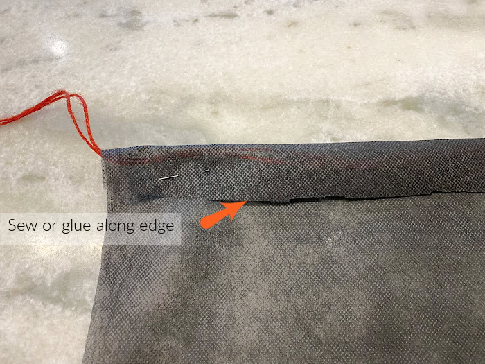 A piece of landscape cloth with the top folded over and pinned.  It is strung through with twine and an arrow points to where it needs to be sewn or glued.