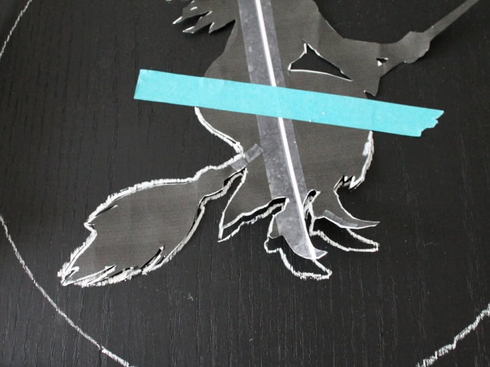 A closeup of the witch being outlined using a white chalk wax pen from Dollar Tree, Part of the template is lifted up so you can see the outline.