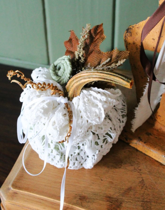 A vintage doily craft pumpkin with a real stem and pumpkin vines. It has a real stem brown leaves and a rolled green ribbon flower. A pretty and neutral vintage fall decor idea for the living room or bedroom.