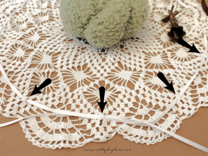 A large doily laying flat with a green pumpkin in the middle. There are arrows pointing to where ribbon was thread through to gather the vintage doily into a pumpkin.