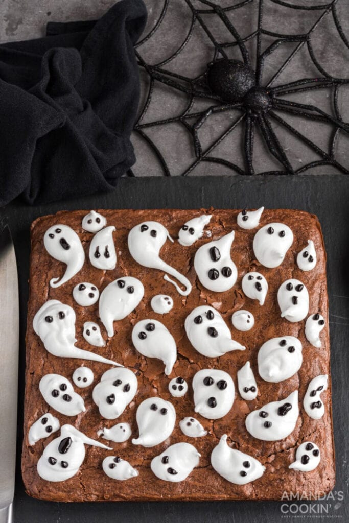 An 8 by 8 slab of homemade Halloween ghost chocolate brownies.  The entire slab is covered in thick chocolate icing with white globs and swirls of white icing on top with little chocolate chip faces.