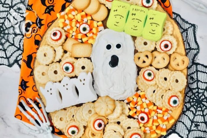 A ghost frosting board, with n icing center in frosting with chocolate eyes and mouth its surrounded by various dippers including marshmallow ghosts and green monster marshmallows, eyeball cookies and small round cookies for dipping.