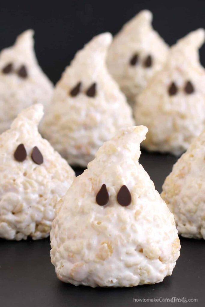 The close up view of several ghost mounds made out of rice krispy treats.  Each ghost has a off kilter pointed top and chocolate chip eyes.