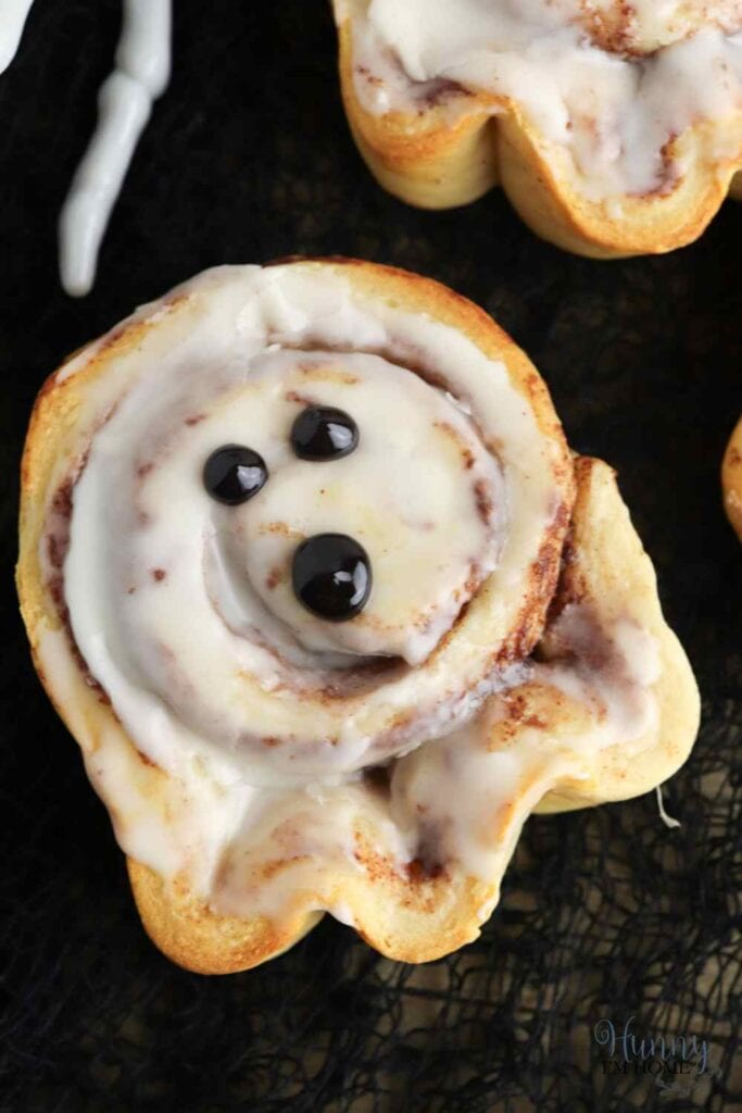 A closeup of a single ghost cinnamon bun decorated in white icing with chocolate chip eyes and mouth.  