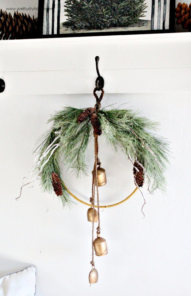 Close-up of a simple Christmas wreath with gold farmhouse bells hanging on a black vintage hook against a white wall. The gold wire and greenery pop against the bright farmhouse background.