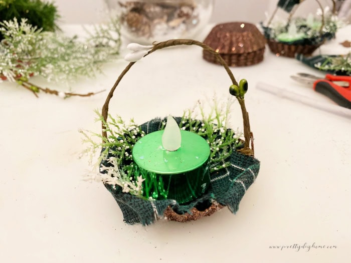 A tartin tin diy Christmas tree ornament almost finished, it has fabric, greenery a handle and a painted glittery tart tin. the back ground shows the remaining pretty glittering and sparkling tart tins and greenery in the background.