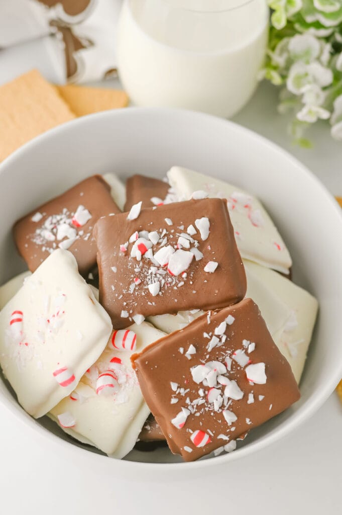 A closeup of a white bowl filled with white chocolate and milk chocolate covered graham wafers that are all sprinkled with peppermint.
