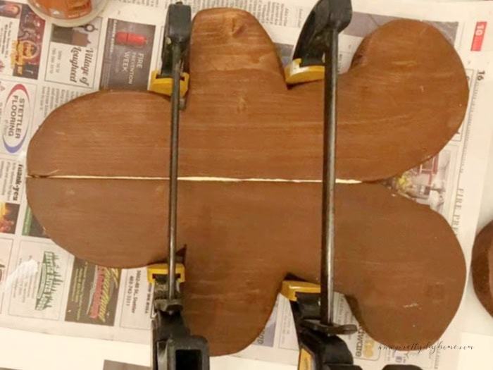 Wooden gingerbread man laying flat on newspaper with glue applied between the two halves and clamps holding the sides in place while the glue dries.