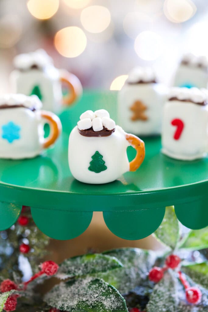 Christmas oreo balls deorated to looks like tiny cups of hot chocolate with pretzel handles, candy designs on each oreo ball front, and topped with mini marshmallows. The little hot chocolate mugs are sitting on a green tray with holly berries and greenery tucked in underneath.