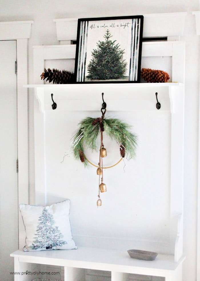 Full view of a minimalist Christmas wreath with gold bells hanging from a black vintage hook on a white hall tree. Above the wreath is tree artwork with pinecones on a shelf, and below is a bench with a white and green Christmas cushion.