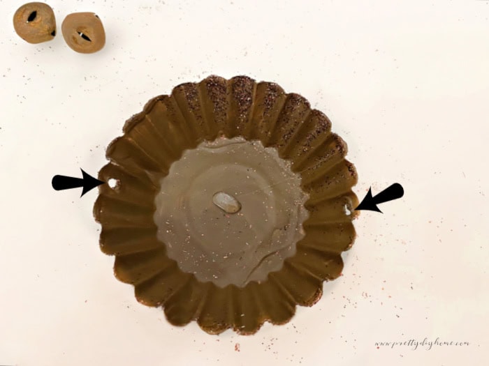 An Inexpensive tart tin painted with two coats of milk chocolate paint and glitter on the outside, and one coat on the inside. There are two arrows pointing to the punched holes used to attach a pretty paper wire hanger while making a DIY Christmas ornament