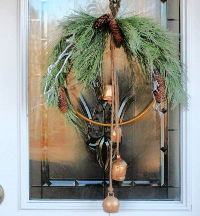 Close up of a simple Christmas wreath with gold farmhouse bells hanging on the front door with cut glass panels.  The wreath is wrapped in gold wire with soft pine greenery and white flocked branches at the top.