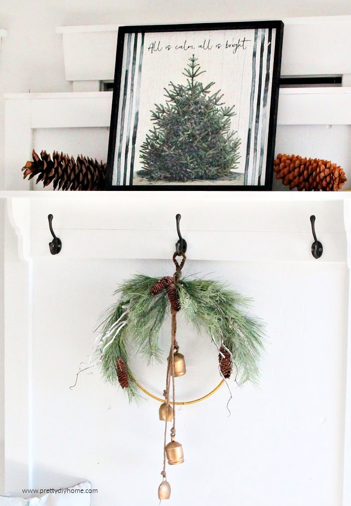 Simple Christmas wreath with cascading gold bells hanging on a white farmhouse hall tree. The wreath sits beneath a shelf with pinecones and greenery.