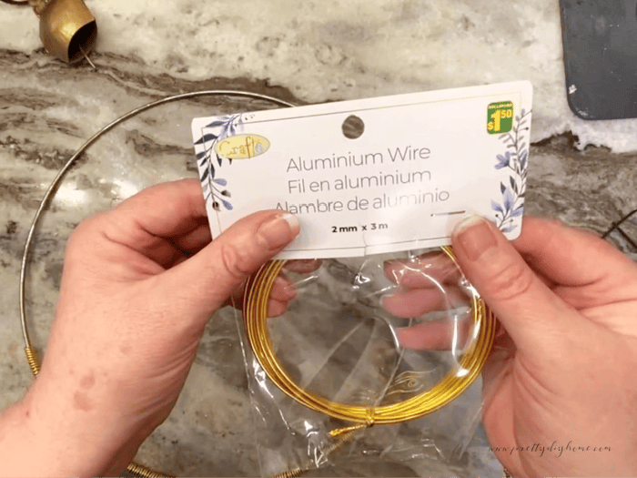 I am holding up a coil of gold aluminum wire used to make a simple Christmas wreath. In the background are the thrifted metal ring, farmhouse bells there is also greenery.