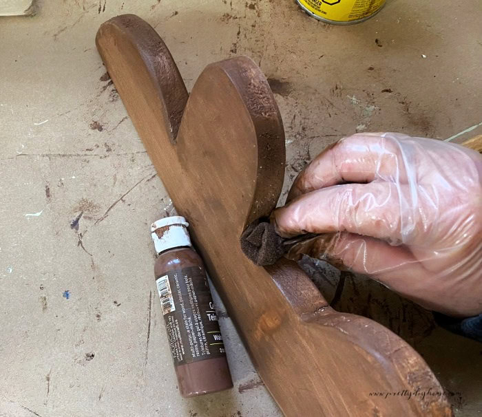 Applying brown wood stain with a rag to half of a wooden gingerbread man lying on its side to cover both the face and edges evenly.