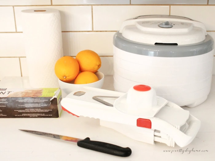 Supplies for drying orange slices with a dehydrator, including fresh oranges, a mandoline slicer, paper towels, and trays.