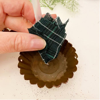 Adding a quarter folded center of a piece of tartan fabric to the center of a brown painted and glittered tart tin, The tart tin has hot glue to hot the center so a hot glue stick is being used to push the fabric in place while the glue is still hot.