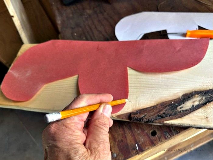 Tracing half of a gingerbread man pattern onto the edge of a 1x6 scrap wood board using a pencil for a DIY wooden gingerbread craft.